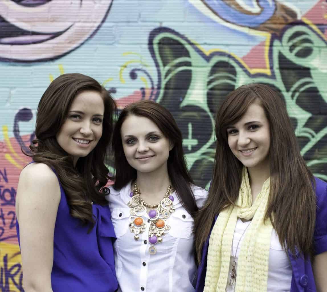 "Three young girls standing outdoors, smiling."